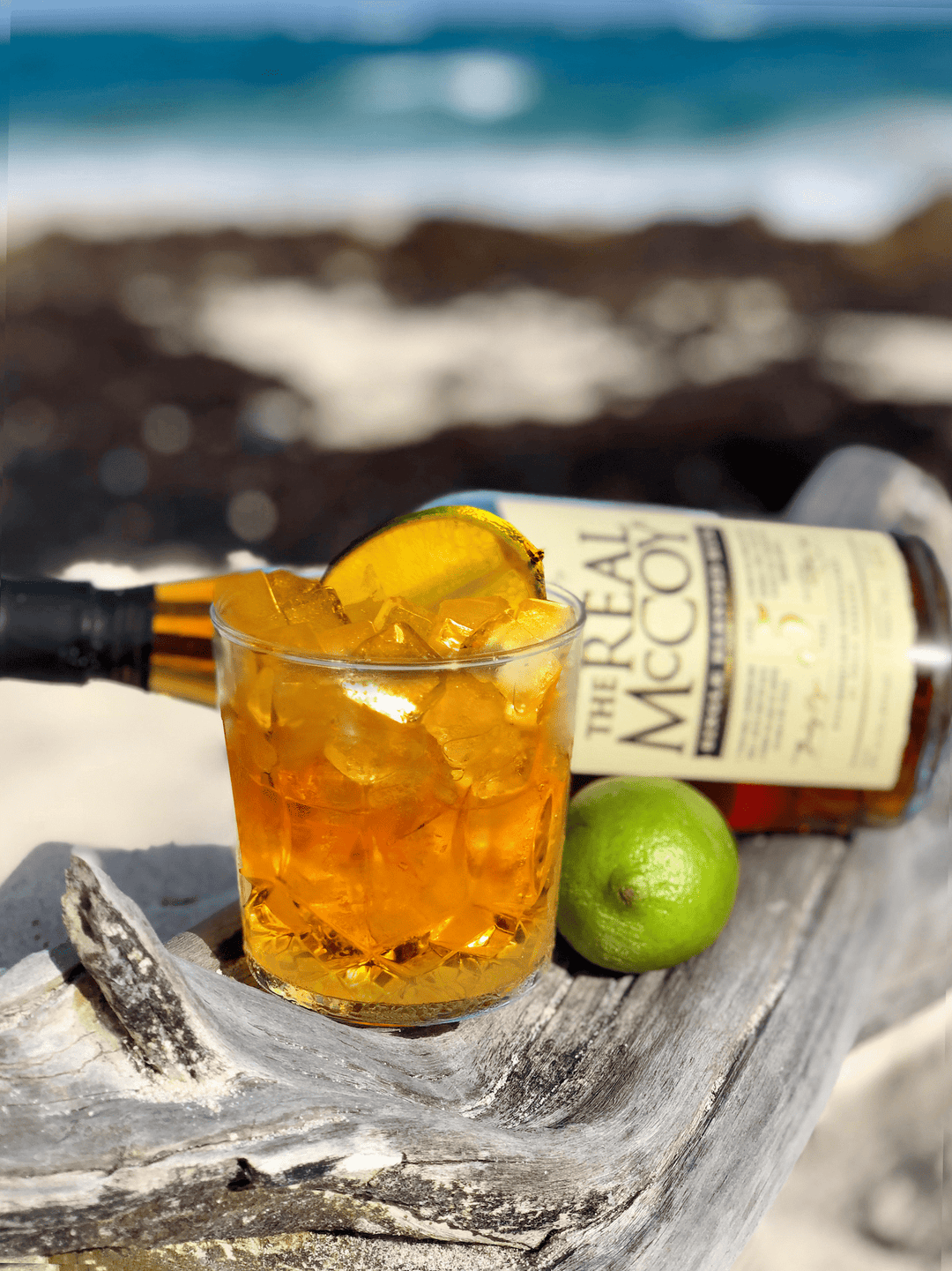 Image of a Corn n Oil cocktail in rocks glass sitting atop a piece of driftwood on the beach in Barbados with a bottle of Real McCoy 5 year aged rum in the background.
