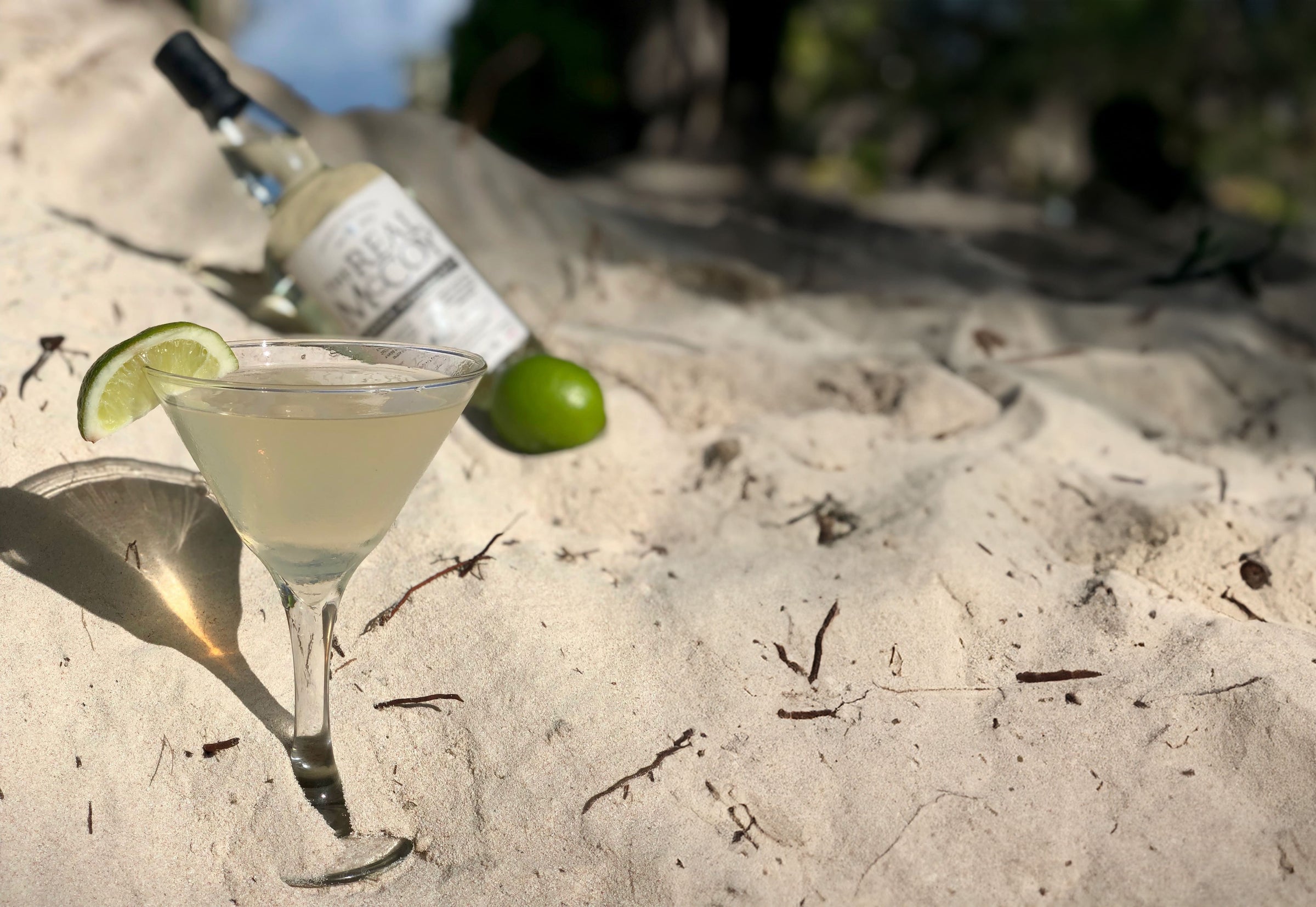 photo of a daiquiri cocktail in a stem glass with a lime wedge sat in the sand with a bottle of Real McCoy 3 year aged rum in the background.