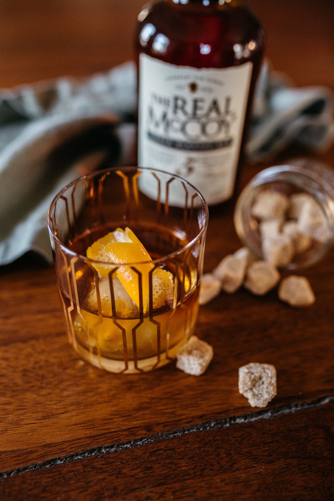 Image of a Real McCoy 5 year aged rum old fashioned cocktail in a rocks with an orange twist garnish. A bottle of Real McCoy 5 Year Aged Rum and some cane sugar cubes are in the background.
