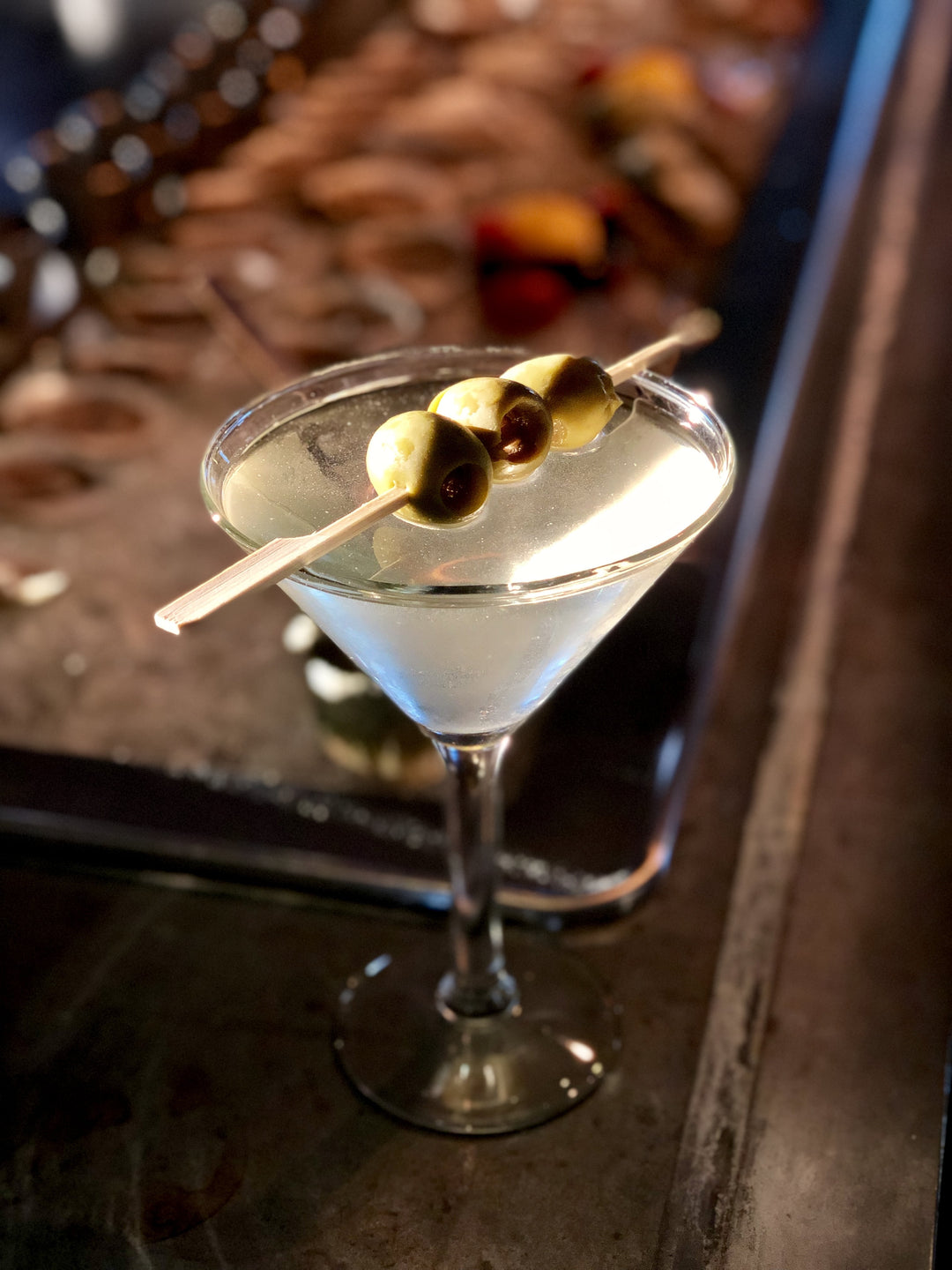 Image of a Dirty Rum Martini garnished with three olives and a tray of oysters on ice in the background.