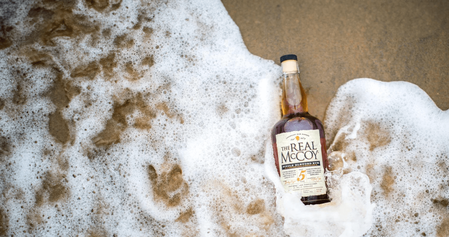 Image of The Real McCoy 5 Year Aged Rum Bottle in the sand with a wave crashing over it