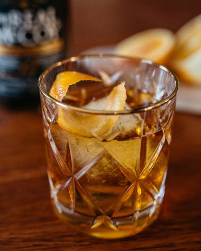 Image of a Real McCoy 12 year aged rum aficionado old fashioned cocktail in a rocks with an orange twist garnish. A bottle of Real McCoy 12 Year Aged Rum and orange slices are in the background.