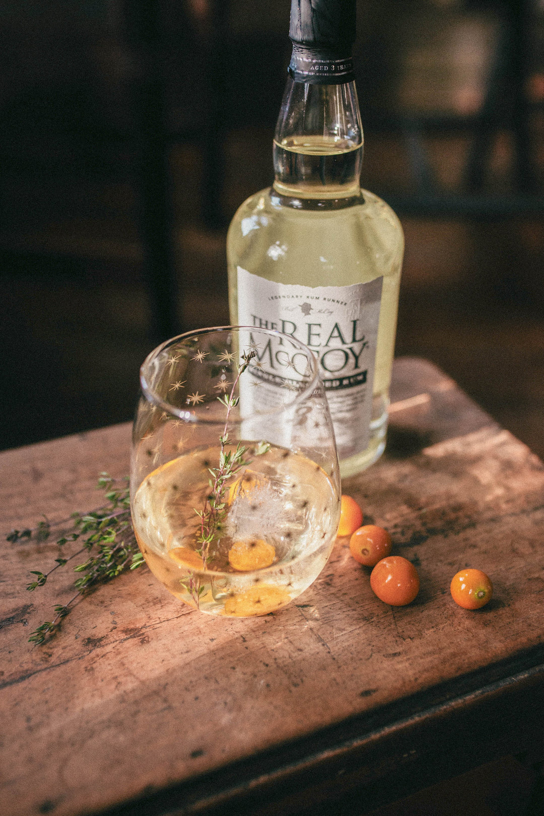 Photo of the white negroni cocktail in a stemless wine glass with orange berries and thyme to garnish sat on a rustic wood table with a bottle of the Real McCoy 3 year aged rum in the background.