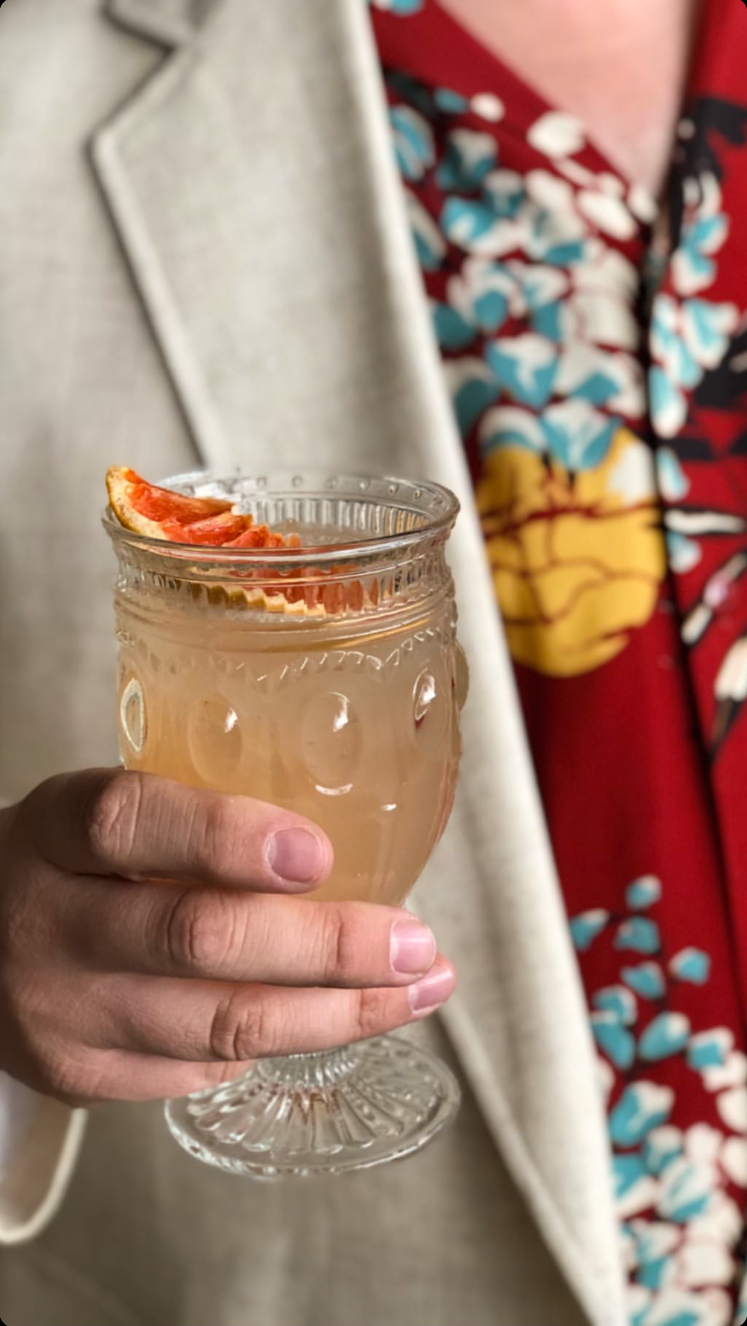 photo of someone with a bright fun patterned shirt holding a Honeysuckle cocktail in vintage stemmed glass with a dehydrated grapefruit slice to garnish