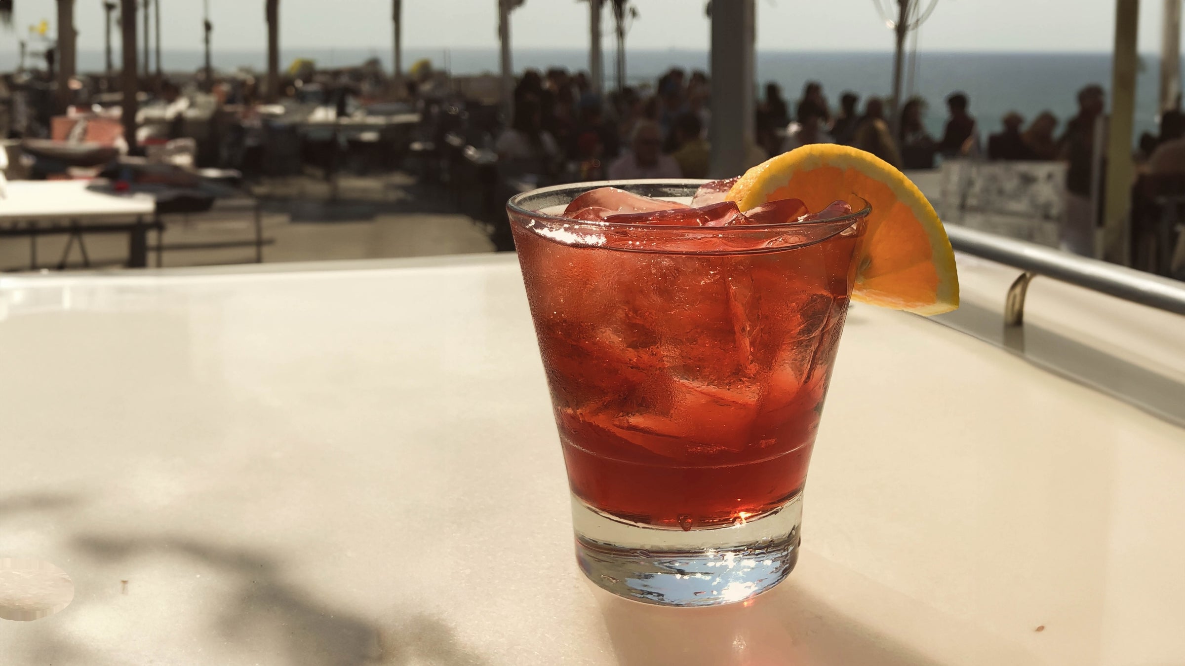Image of a bright red Negroni Paradiso cocktail with an orange peel garnish on a bartop at a rooftop, ocean view bar.