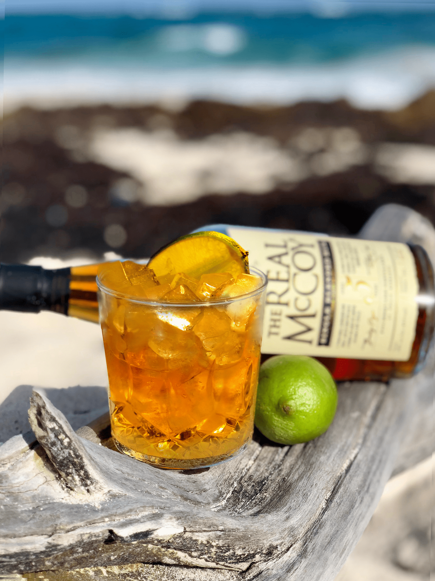 Image of a Corn n Oil cocktail in rocks glass sitting atop a piece of driftwood on the beach in Barbados with a bottle of Real McCoy 5 year aged rum in the background.